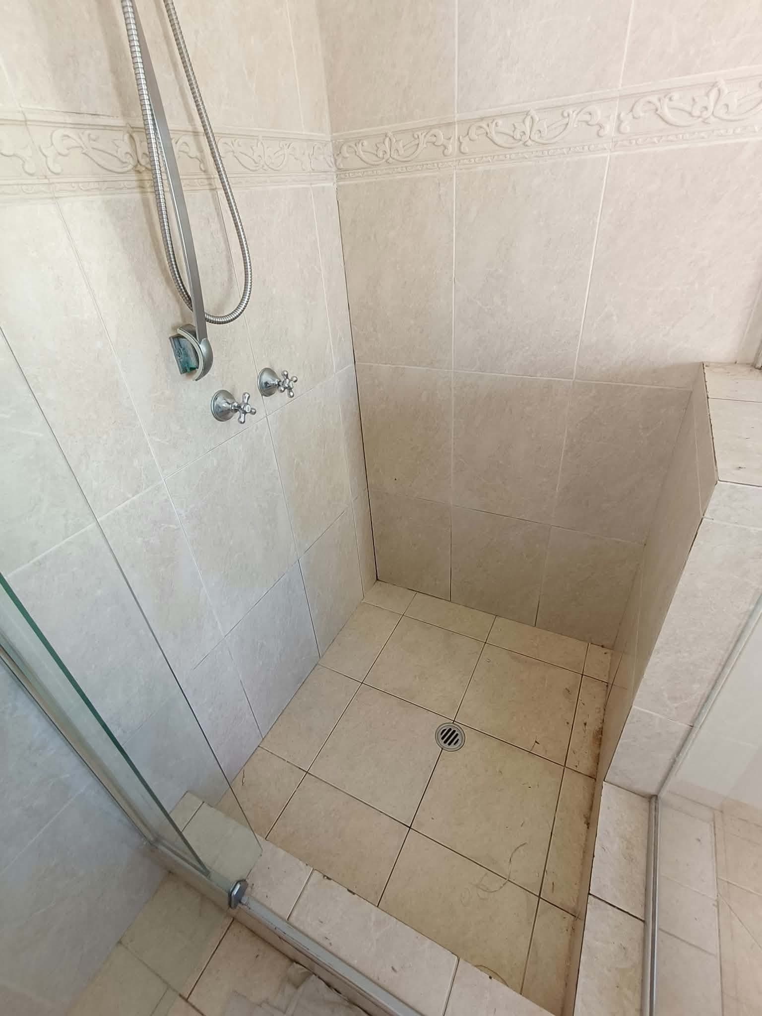 Beige stone-look shower base before regrouting, with patchy discoloured grout across the floor and a darkened, debris-filled joint at the wall