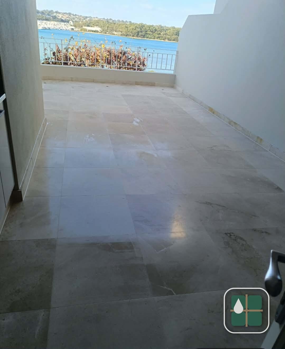 Large stone-tiled balcony terrace overlooking water, with wide-format pale tiles, tight grout lines and a sealed junction at the balustrade wall.